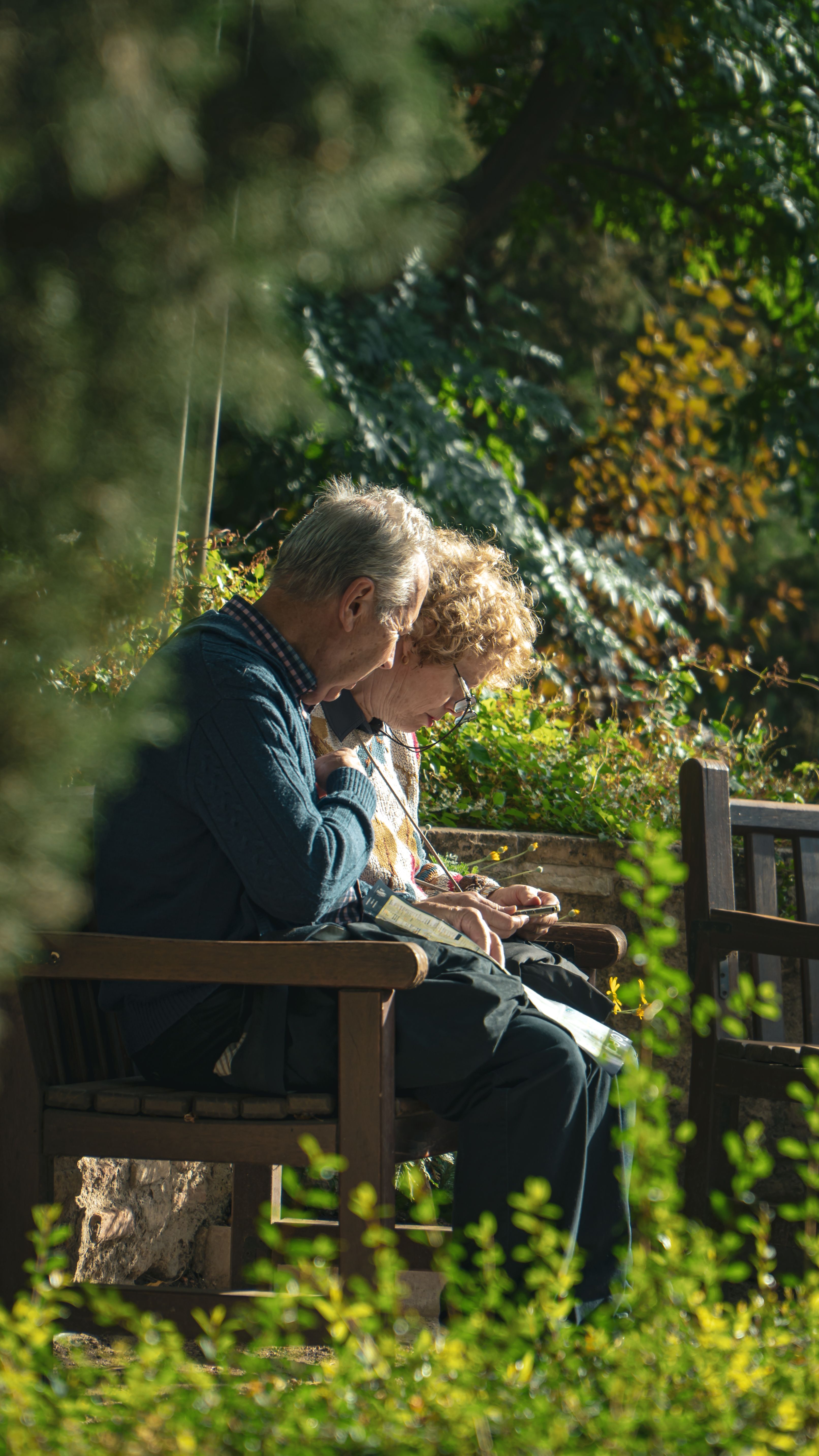 An older couple sit close together on a garden bench, reading something in the afternoon sun.