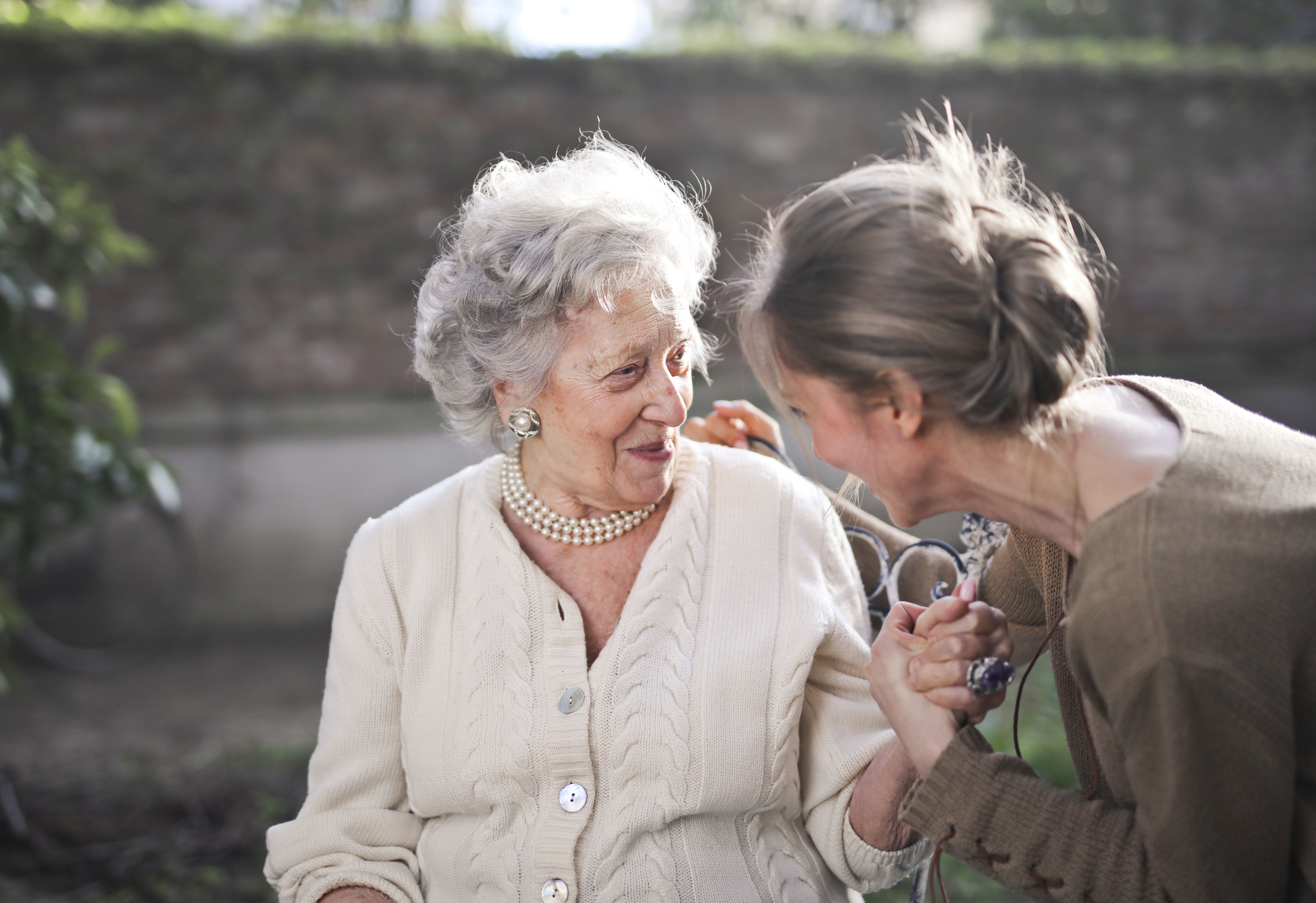 An older woman and a younger woman lean close together outdoors, talking and smiling.