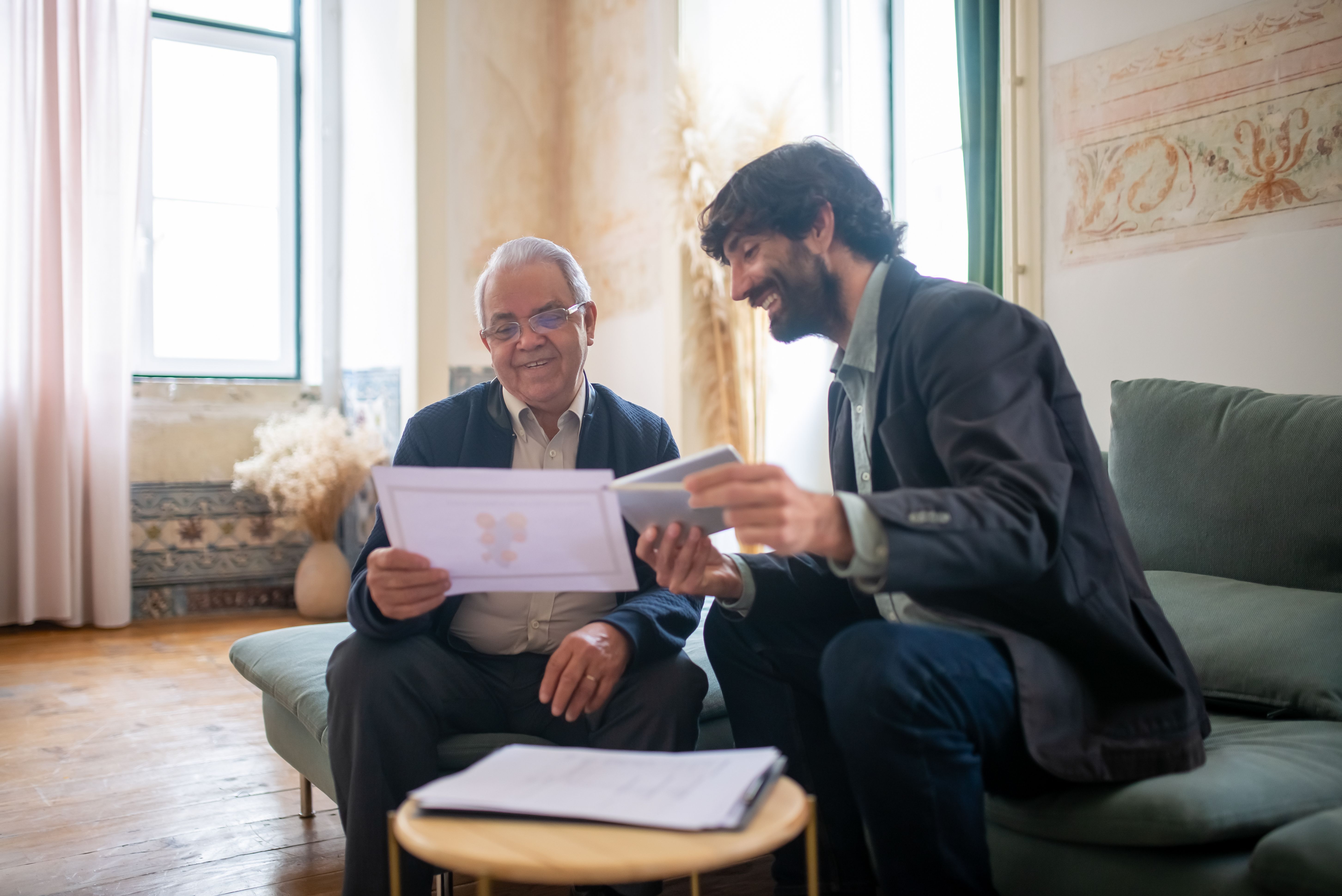 An older man and a younger man sit together on a sofa, going through paperwork and smiling.