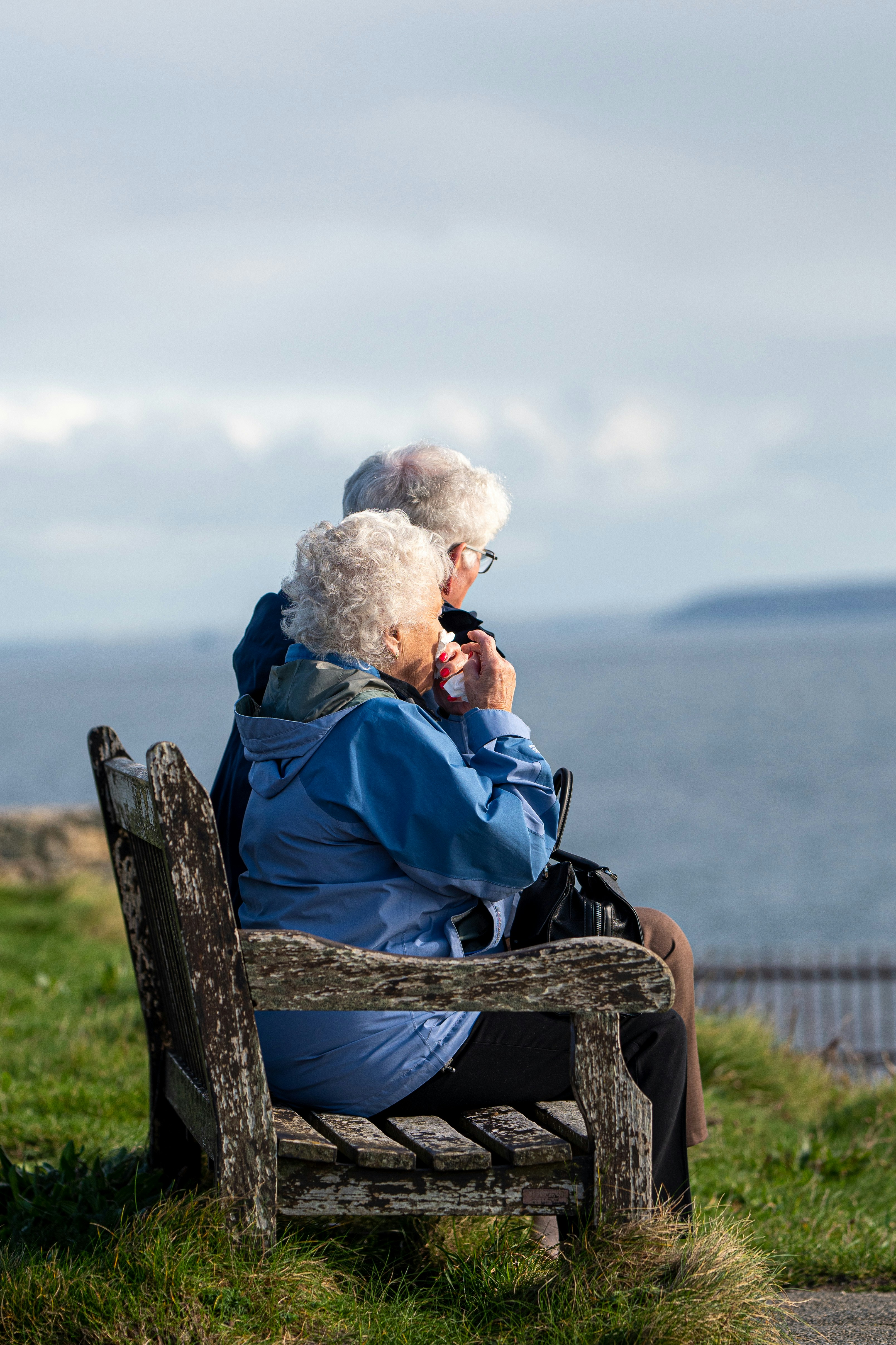 An older couple sit close together on a weathered bench, looking out over the sea.